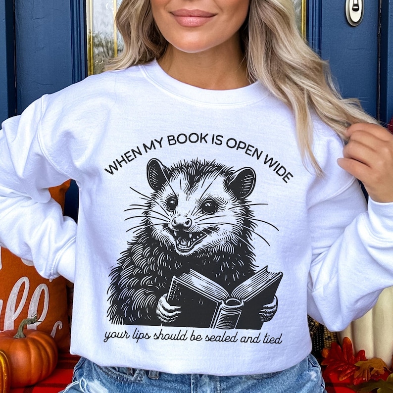 May include: White sweatshirt with a black and white illustration of an opossum reading a book. The text on the sweatshirt reads "When my book is open wide your lips should be sealed and tied."