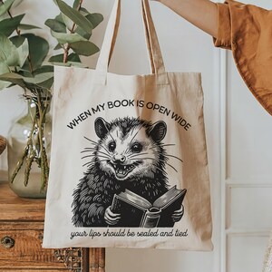 May include: A beige tote bag with a black and white illustration of an opossum holding a book. The text on the bag reads "When my book is open wide your lips should be sealed and tied."