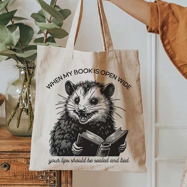 May include: A beige tote bag with a black and white illustration of an opossum holding a book. The text on the bag reads "When my book is open wide your lips should be sealed and tied."