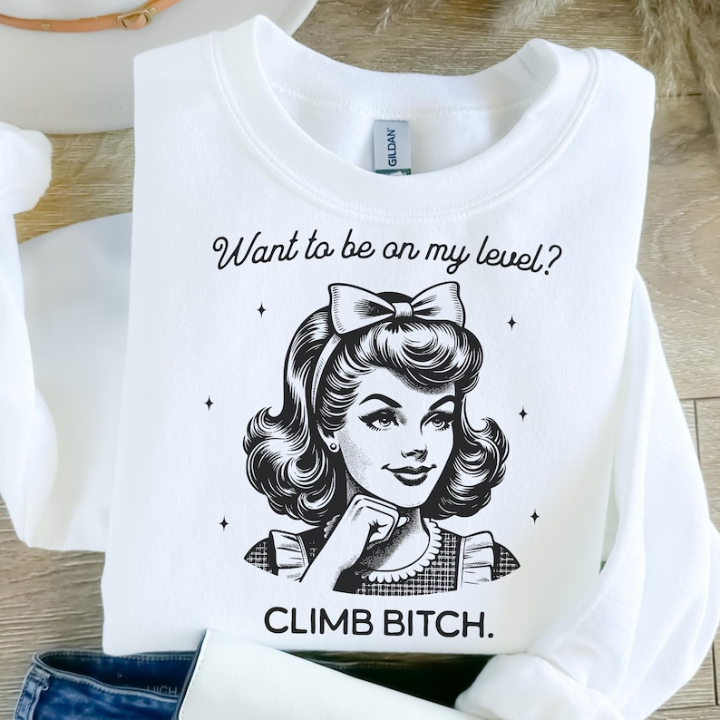 May include: A white long-sleeve sweatshirt featuring a black graphic illustration of a person with a bow in their hair. The text above reads "Want to be on my level?" and the text below reads "CLIMB BITCH." The garment has a crew neckline.