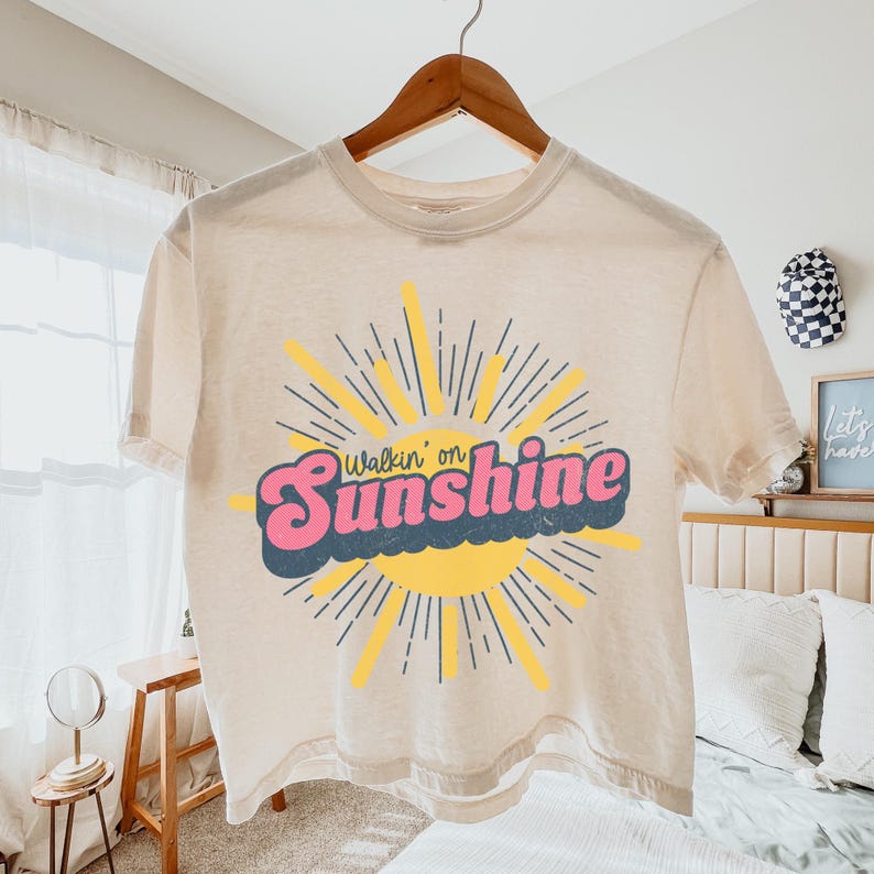 May include: A cream-colored short-sleeve t-shirt featuring a graphic of a yellow sun with radiating lines. The text "Walkin' on Sunshine" is printed across the center in a bold, pink, retro-style font with a dark blue shadow effect.