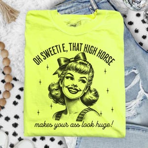 May include: A neon yellow t-shirt featuring a black graphic illustration of a smiling person with a bow in their hair. The text reads: OH SWEETIE, THAT HIGH HORSE makes your ass look huge! The design includes small star accents surrounding the portrait.