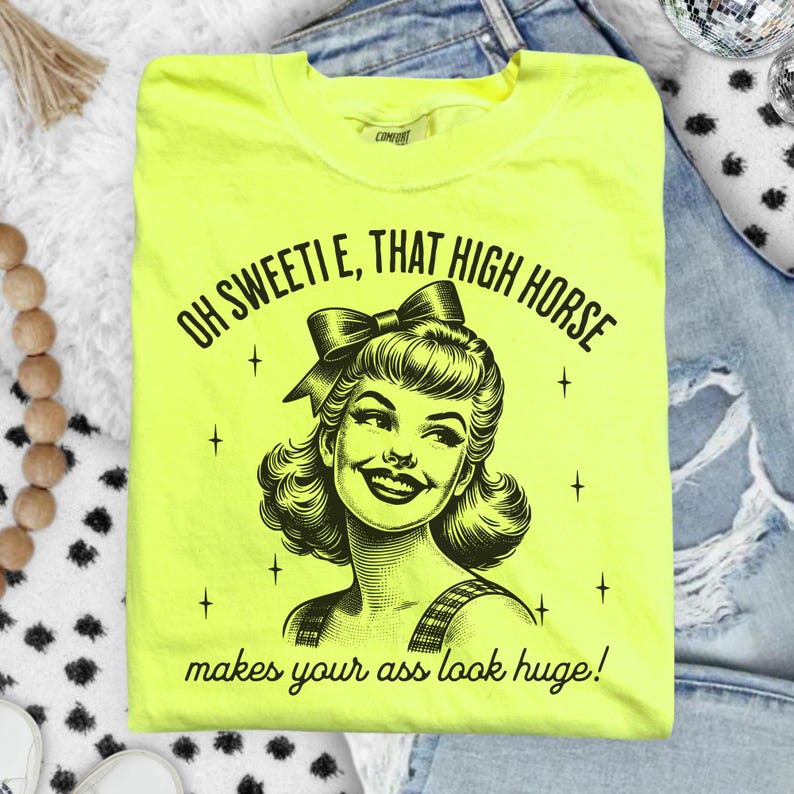 May include: A neon yellow t-shirt featuring a black graphic illustration of a smiling person with a bow in their hair. The text reads: OH SWEETIE, THAT HIGH HORSE makes your ass look huge! The design includes small star accents surrounding the portrait.