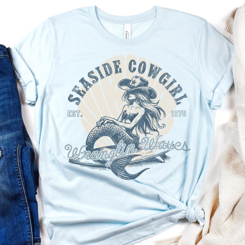 K&ouml;nnte beinhalten: Hellblaues T-Shirt mit einem Grafikdruck einer Meerjungfrau, die einen Cowboyhut tr&auml;gt und auf einer Welle reitet. Der Text "Seasde Cowgirl Est. 1975 Wranglin' Waves" ist auf dem Shirt aufgedruckt.
