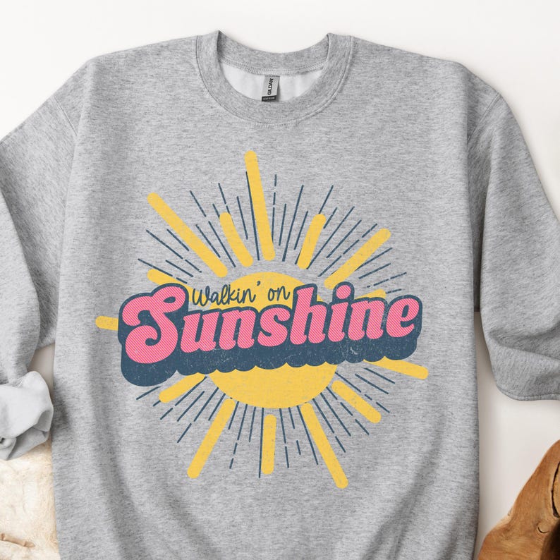 May include: A gray sweatshirt with a retro-style graphic of a sun with rays and the text "Walkin' on Sunshine" in pink and blue letters.