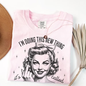 May include: A light pink t-shirt featuring a black graphic print of a person with a bow in their hair, giving the middle finger. The text reads, "I'M DOING THIS NEW THING it's called" above and below the graphic. A delicate necklace rests on the shirt.