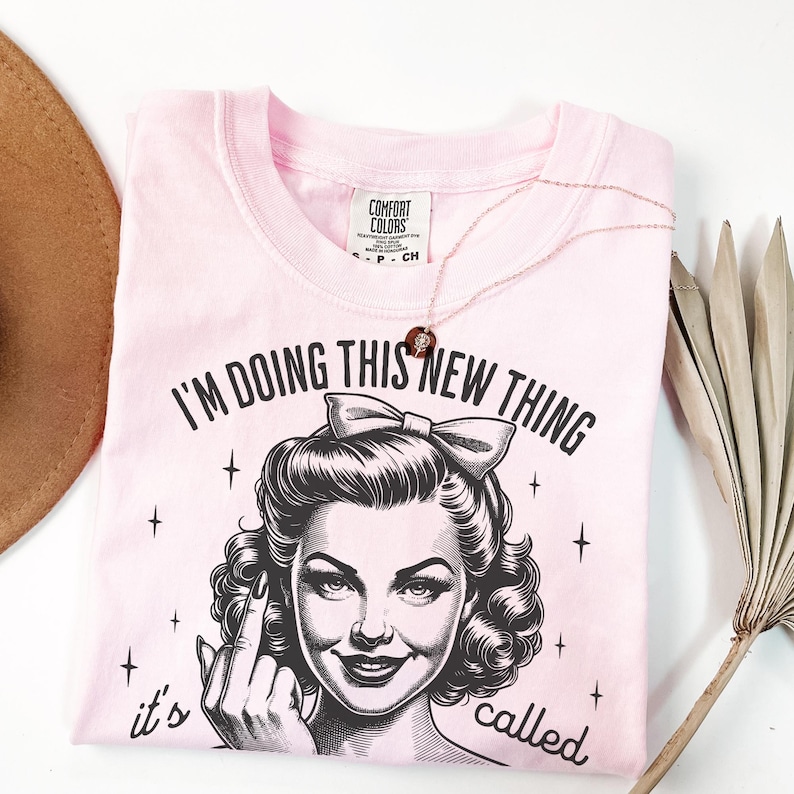 May include: A light pink t-shirt featuring a black graphic print of a person with a bow in their hair, giving the middle finger. The text reads, "I'M DOING THIS NEW THING it's called" above and below the graphic. A delicate necklace rests on the shirt.