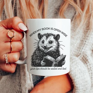 May include: White ceramic mug with a black and white illustration of an opossum reading a book. The text on the mug reads "When my book is open wide your lips should be sealed and tied."