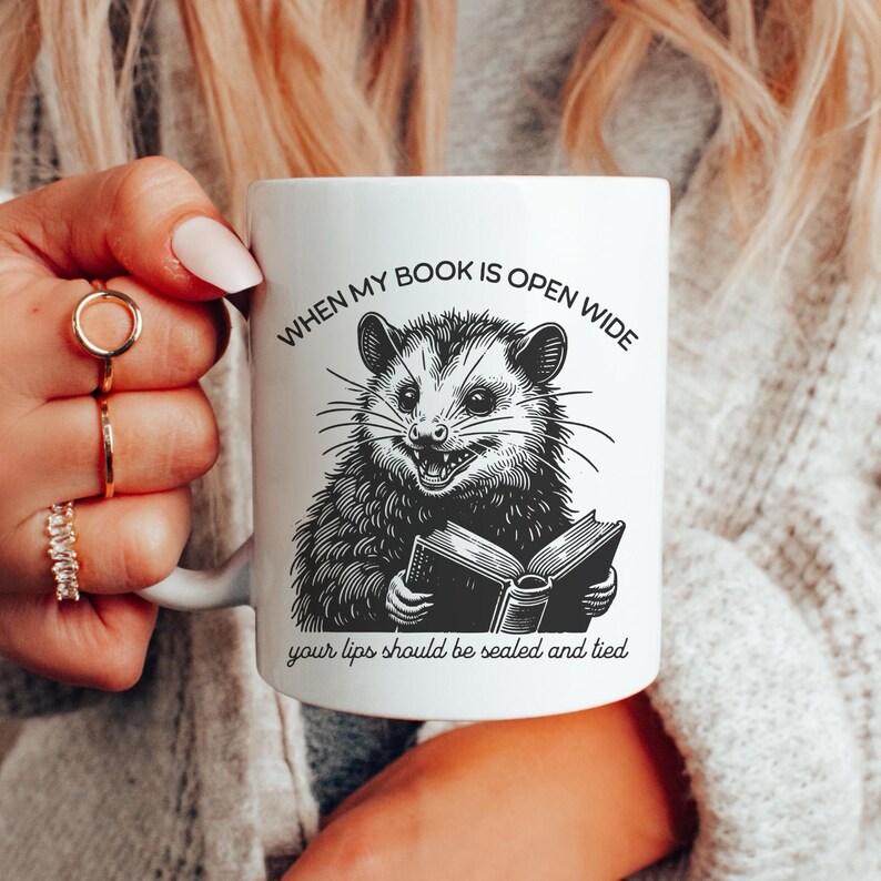 May include: White ceramic mug with a black and white illustration of an opossum reading a book. The text on the mug reads "When my book is open wide your lips should be sealed and tied."