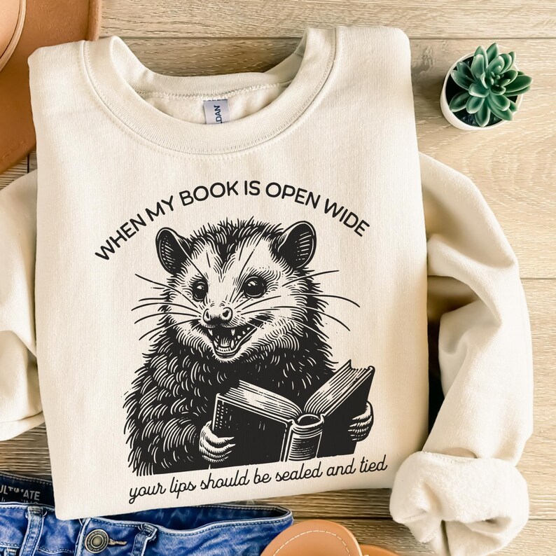 May include: A cream-colored sweatshirt with a black and white illustration of an opossum reading a book. The text "WHEN MY BOOK IS OPEN WIDE your lips should be sealed and tied" is printed on the sweatshirt.