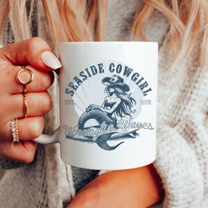 K&ouml;nnte beinhalten: Eine wei&szlig;e Keramiktasse mit einer blauen und wei&szlig;en Illustration einer Meerjungfrau, die einen Cowboyhut tr&auml;gt und auf einem Surfbrett reitet. Der Text "Seasde Cowgirl Est. 1975 Wranglin' Waves" ist auf die Tasse gedruckt.