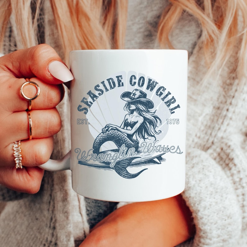 K&ouml;nnte beinhalten: Eine wei&szlig;e Keramiktasse mit einer blauen und wei&szlig;en Illustration einer Meerjungfrau, die einen Cowboyhut tr&auml;gt und auf einem Surfbrett reitet. Der Text "Seasde Cowgirl Est. 1975 Wranglin' Waves" ist auf die Tasse gedruckt.