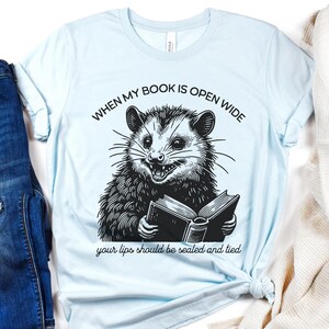 May include: Light blue t-shirt with a black and white illustration of a possum holding a book. The text on the shirt reads "When my book is open wide your lips should be sealed and tied."