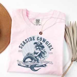 K&ouml;nnte beinhalten: Pinkes T-Shirt mit einer blauen Grafik einer Meerjungfrau, die einen Cowboyhut tr&auml;gt und auf einem Surfbrett reitet. Die Grafik lautet "Seasside Cowgirl Est. 1975 Wranglin' Waves".