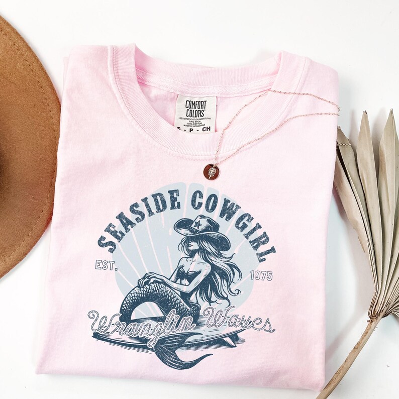 K&ouml;nnte beinhalten: Pinkes T-Shirt mit einer blauen Grafik einer Meerjungfrau, die einen Cowboyhut tr&auml;gt und auf einem Surfbrett reitet. Die Grafik lautet "Seasside Cowgirl Est. 1975 Wranglin' Waves".