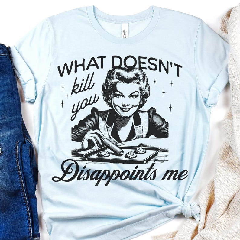 May include: A light blue t-shirt featuring a black graphic illustration of a person with a vintage hairstyle using a rolling pin on a baking sheet with cookies. The text reads: WHAT DOESN'T kill you Disappoints me, with small star accents.