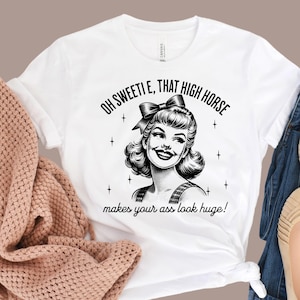 May include: A white t-shirt featuring a black graphic illustration of a smiling person with a bow in their hair, surrounded by small star icons. The text reads, "OH SWEETIE, THAT HIGH HORSE makes your ass look huge!" in a bold, black, sans-serif font.
