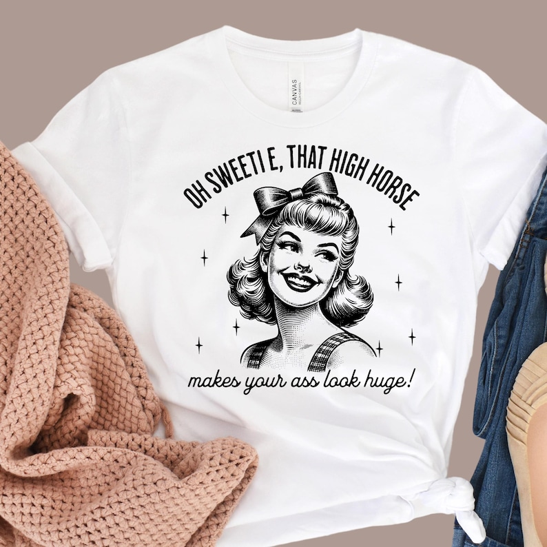 May include: A white t-shirt featuring a black graphic illustration of a smiling person with a bow in their hair, surrounded by small star icons. The text reads, "OH SWEETIE, THAT HIGH HORSE makes your ass look huge!" in a bold, black, sans-serif font.