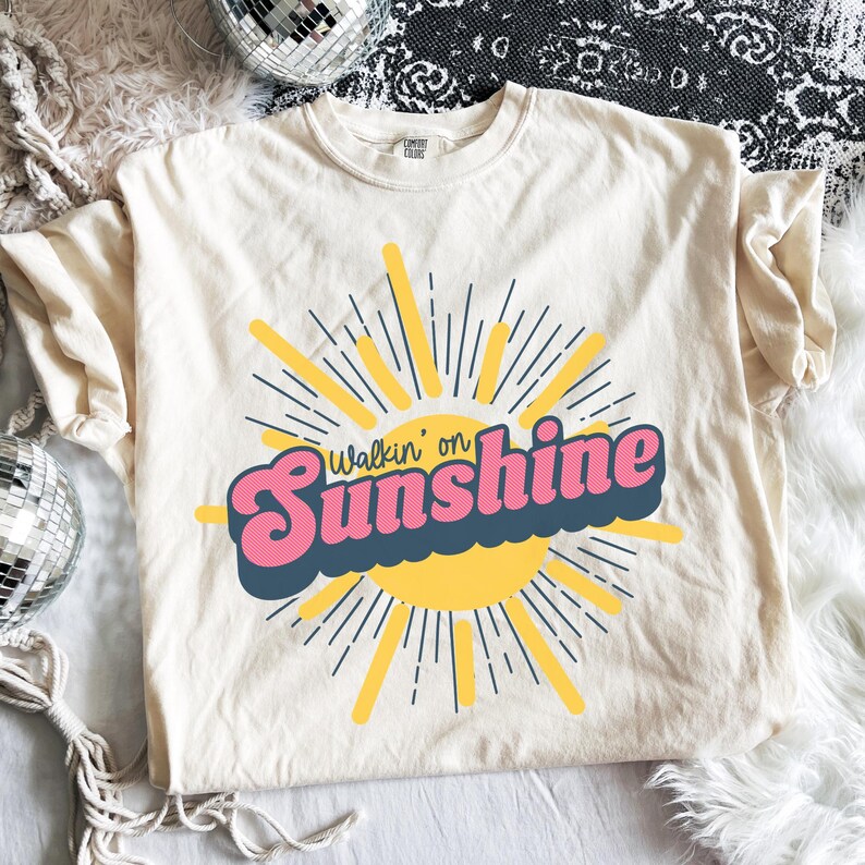 May include: A cream-colored short-sleeve t-shirt featuring a graphic of a yellow sun with radiating lines. The text "Walkin' on Sunshine" is printed across the center in a bold, pink, retro-style font with a dark blue shadow effect.