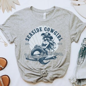 K&ouml;nnte beinhalten: Graues T-Shirt mit einem blauen Motiv einer Meerjungfrau, die einen Cowboyhut tr&auml;gt und auf einem Surfbrett reitet. Das Motiv enth&auml;lt den Text "Seashore Cowgirl Est. 1975 Wranglin' Waves".