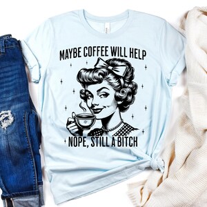 May include: A light blue t-shirt featuring a black graphic illustration of a person with a bow in their hair holding a coffee cup. The text above reads MAYBE COFFEE WILL HELP and the text below reads NOPE, STILL A BITCH, surrounded by small stars.