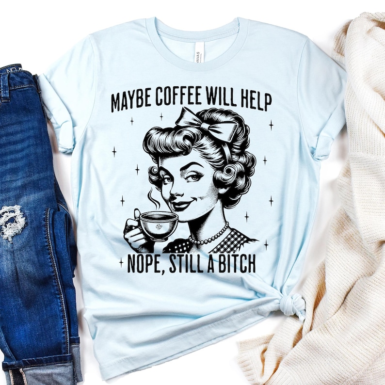 May include: A light blue t-shirt featuring a black graphic illustration of a person with a bow in their hair holding a coffee cup. The text above reads MAYBE COFFEE WILL HELP and the text below reads NOPE, STILL A BITCH, surrounded by small stars.