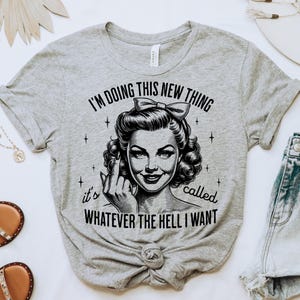May include: A heather grey t-shirt featuring a black graphic of a person with a bow in their hair, giving the middle finger. The text reads: I'M DOING THIS NEW THING it's called WHATEVER THE HELL I WANT. The design includes small star accents.