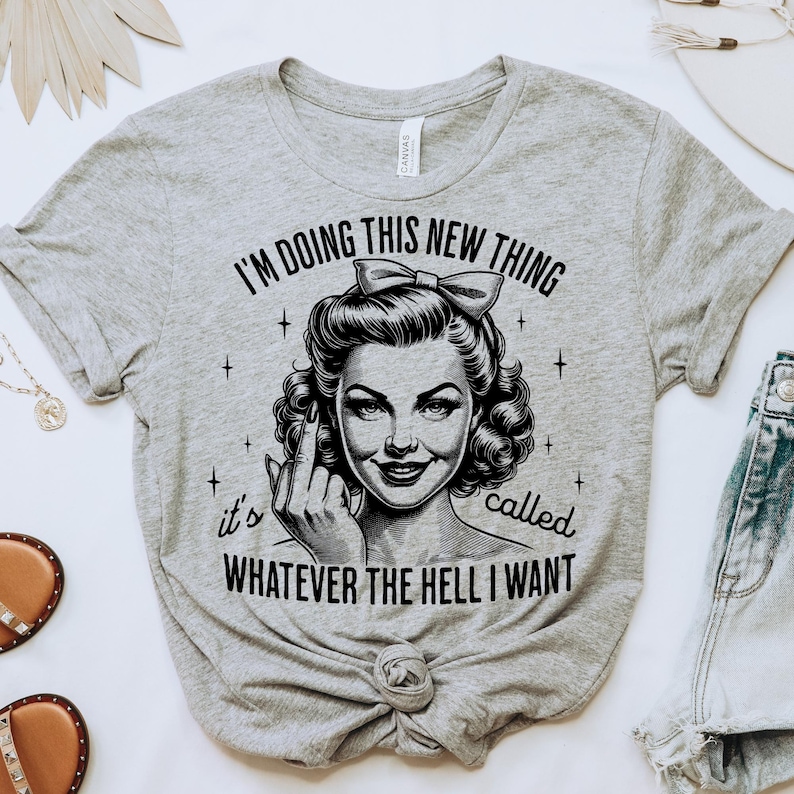 May include: A heather grey t-shirt featuring a black graphic of a person with a bow in their hair, giving the middle finger. The text reads: I'M DOING THIS NEW THING it's called WHATEVER THE HELL I WANT. The design includes small star accents.