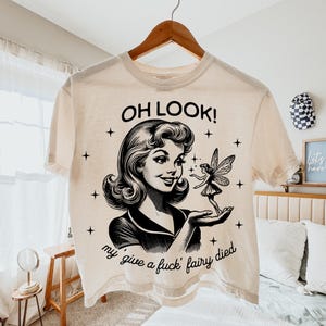 May include: A cream-coloured short-sleeve t-shirt featuring a black graphic print of a woman looking at a small fairy in her hand. The text reads OH LOOK! above the woman and my give a fuck fairy died below her, surrounded by small star icons.