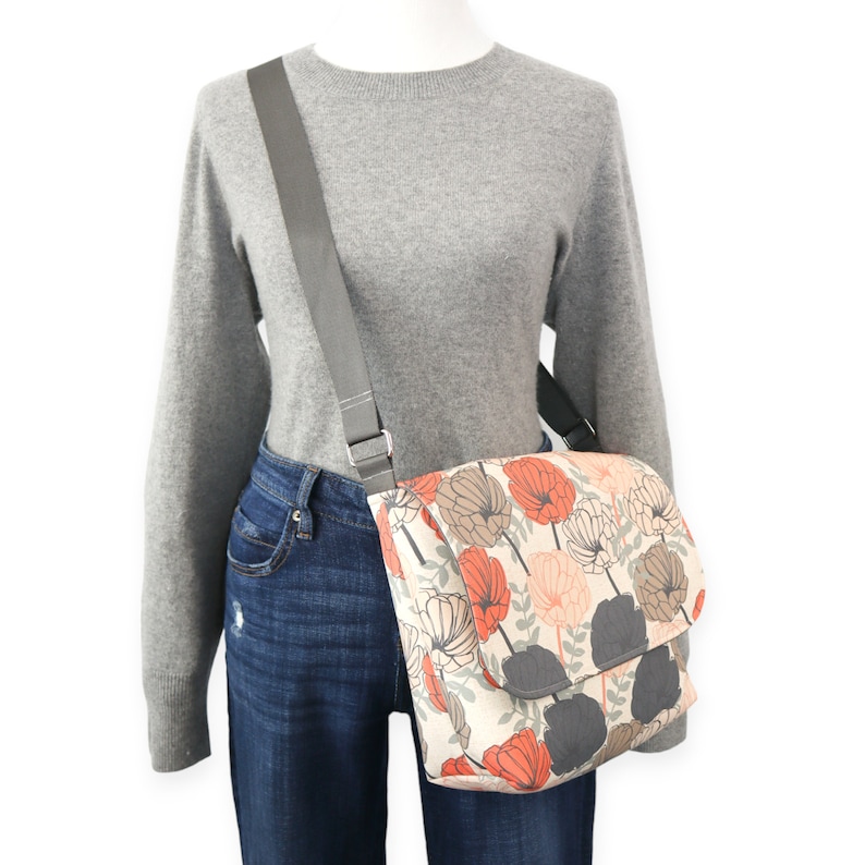 May include: A gray canvas messenger bag with a floral print featuring orange, pink, and brown flowers. The bag has a gray adjustable strap.