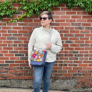 May include: A woman wearing a light beige jacket and blue jeans is standing in front of a brick wall. She is wearing sunglasses and carrying a blue and floral patterned crossbody bag.