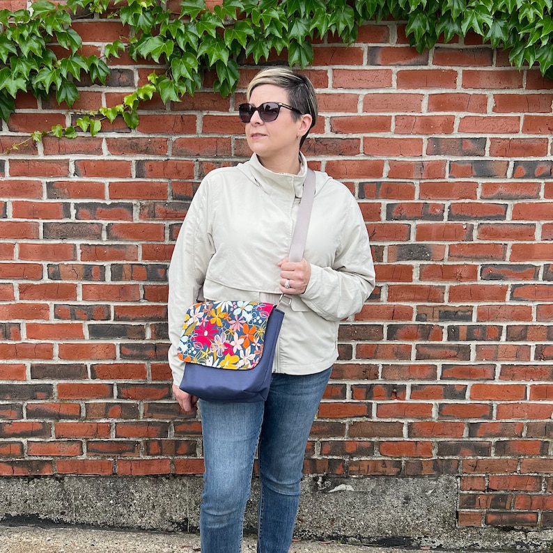 May include: A woman wearing a light beige jacket and blue jeans is standing in front of a brick wall. She is wearing sunglasses and carrying a blue and floral patterned crossbody bag.