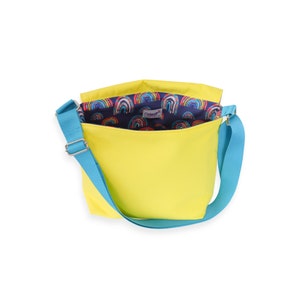 May include: A yellow canvas crossbody bag with a blue strap. The bag is open and the inside lining is navy blue with a rainbow pattern.