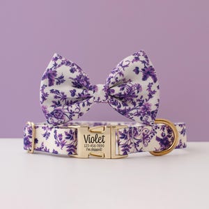 May include: A dog collar and bow tie set with a floral pattern in white and purple. The collar has a gold-colored buckle with the name "Violet" and a phone number. The bow tie is attached to the collar.