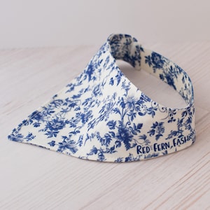 May include: A white visor with a blue floral pattern. The visor has a curved brim and a band that goes around the head. The text "RED FERN, FASHION" is printed on the side of the visor.