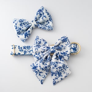May include: Two matching pet accessories: a bow tie and a collar with a bow. Both are white with a blue floral pattern. The collar has a gold-colored metal buckle and a D-ring. The bow tie is separate.