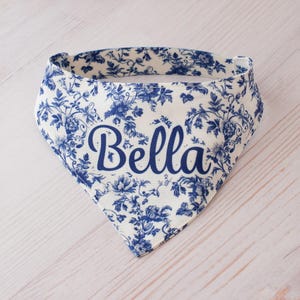 May include: A white and blue floral patterned dog bandana with the name "Bella" in a cursive font. The bandana is triangular in shape and features a classic toile design. The fabric is a light color with blue floral accents.