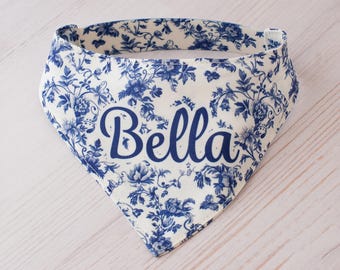May include: A white and blue floral patterned dog bandana with the name "Bella" in a cursive font. The bandana is triangular in shape and features a classic toile design. The fabric is a light color with blue floral accents.