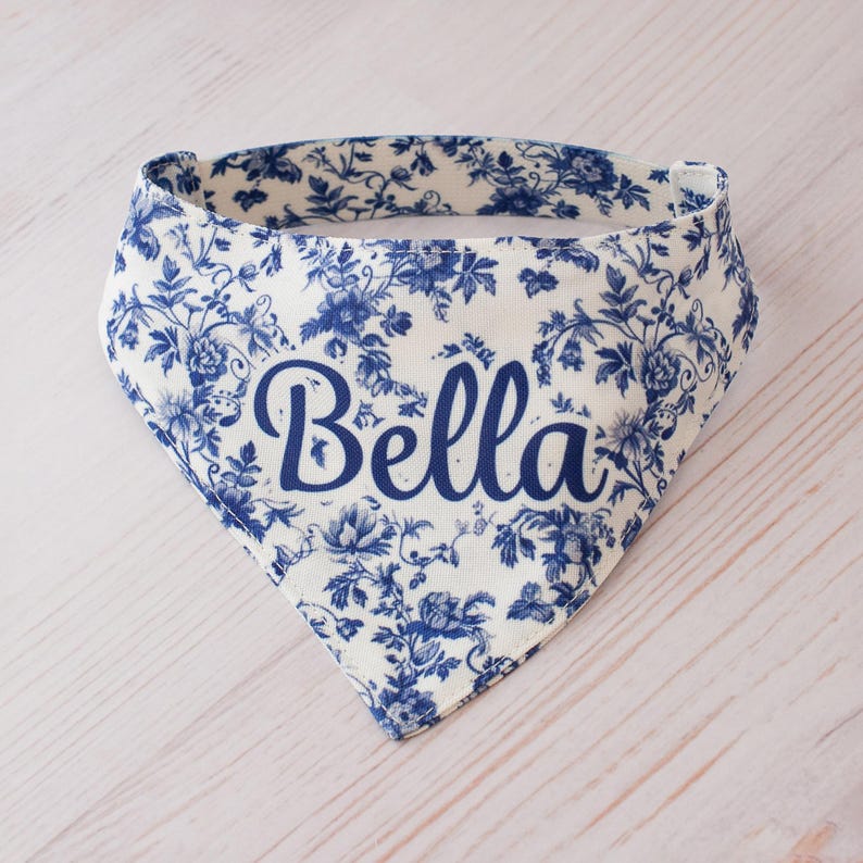 May include: A white and blue floral patterned dog bandana with the name "Bella" in a cursive font. The bandana is triangular in shape and features a classic toile design. The fabric is a light color with blue floral accents.