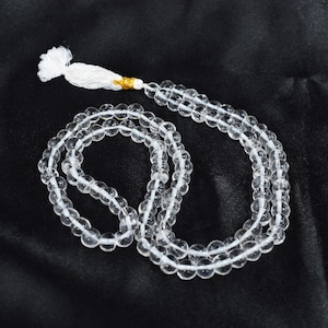 Può includere: Una collana di perline sferiche trasparenti disposte in un motivo circolare, con una nappa bianca a un'estremità fissata da una piccola fascia color oro. Le perline sono traslucide e di dimensioni uniformi, creando un anello continuo di sfere.