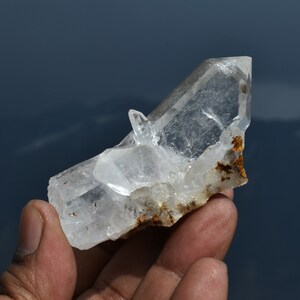 Puede incluir: Un racimo de cristales de cuarzo claro y translúcido que presenta una punta principal alargada y prominente con un cristal secundario más pequeño que sobresale del lado. La base del espécimen mineral muestra inclusiones naturales de óxido de hierro marrón.
