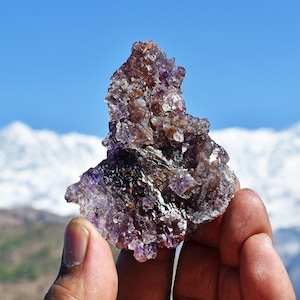 May include: A raw, jagged mineral specimen featuring a deep purple and brown crystalline structure with translucent facets. The cluster displays a rough, uneven surface with varying shades of violet and dark earthy tones throughout the formation.