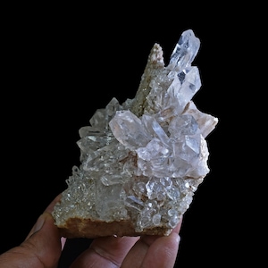 Può includere: Un gruppo di cristalli di quarzo trasparenti su una roccia marrone. I cristalli sono affilati e appuntiti e sono disposti in uno schema casuale. La roccia è ruvida e testurizzata.