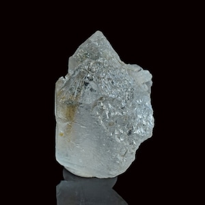 May include: A translucent, colourless quartz crystal cluster with a pointed termination and multifaceted, textured surfaces. The mineral specimen displays internal fractures and a small patch of brownish inclusion near the base, set against a black background.
