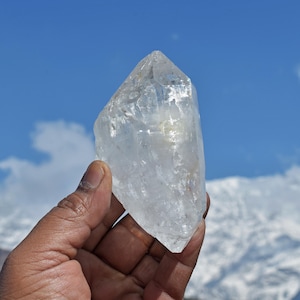 May include: A clear, naturally formed quartz crystal point with a hexagonal shape and multifaceted surface. The crystal is transparent with visible internal inclusions and fractures, showcasing a sharp, pointed termination at the top.