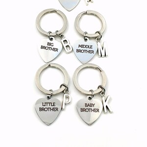 May include: Silver heart-shaped keychains with the words "Brother", "Big Brother", "Middle Brother", "Little Brother", and "Baby Brother". Each keychain has a silver letter charm. The keychains are on a white background.