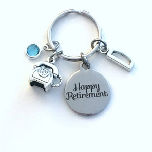Könnte beinhalten: Silberfarbener Schlüsselanhänger mit einem runden Anhänger mit der Gravur "Happy Retirement". Der Schlüsselanhänger enthält einen kleinen Telefonanhänger, einen blauen Edelstein und einen rechteckigen Anhänger. Schlichtes und elegantes Design, als Geschenk zur Rente.