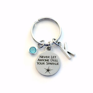 May include: Silver keychain with a round charm that says "Never let anyone dull your sparkle" with a star design. The keychain also has a small blue crystal charm and a silver letter K charm.