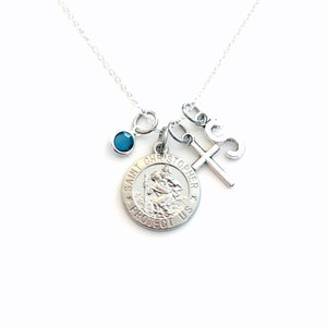 Puede incluir: Collar de plata con una medalla de San Cristóbal, un dije de cruz, un dije con el número 3 y una piedra preciosa azul verdosa.