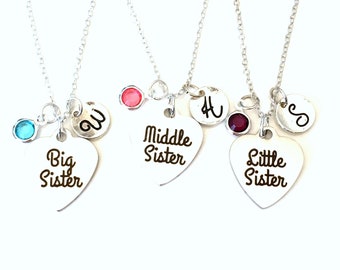 May include: Three silver necklaces with heart-shaped pendants. Each pendant has the words "Big Sister", "Middle Sister", and "Little Sister" printed on them. Each necklace has a different coloured gem and a letter charm.